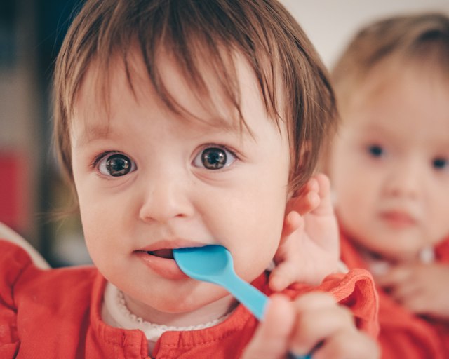 Zwei Kleinkinder essen gemeinsam, eines hat einen Löffel im Mund