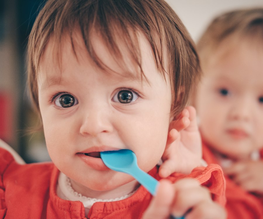 Zwei Kleinkinder essen gemeinsam, eines hat einen Löffel im Mund