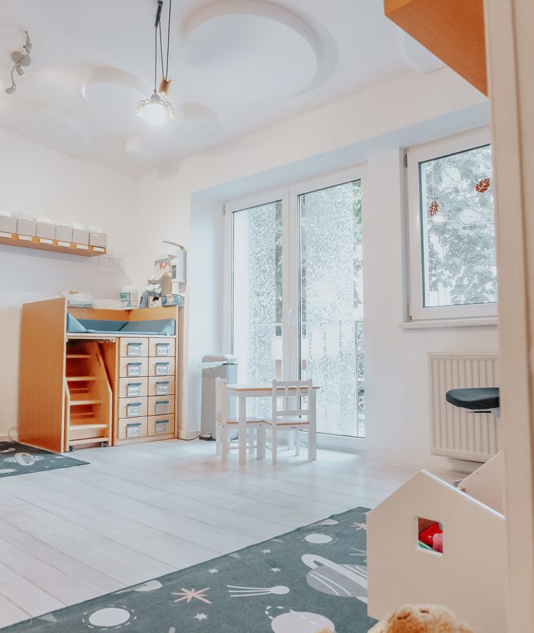 Heller Gruppenraum mit Fensterfront, Regalen und Spielteppich
